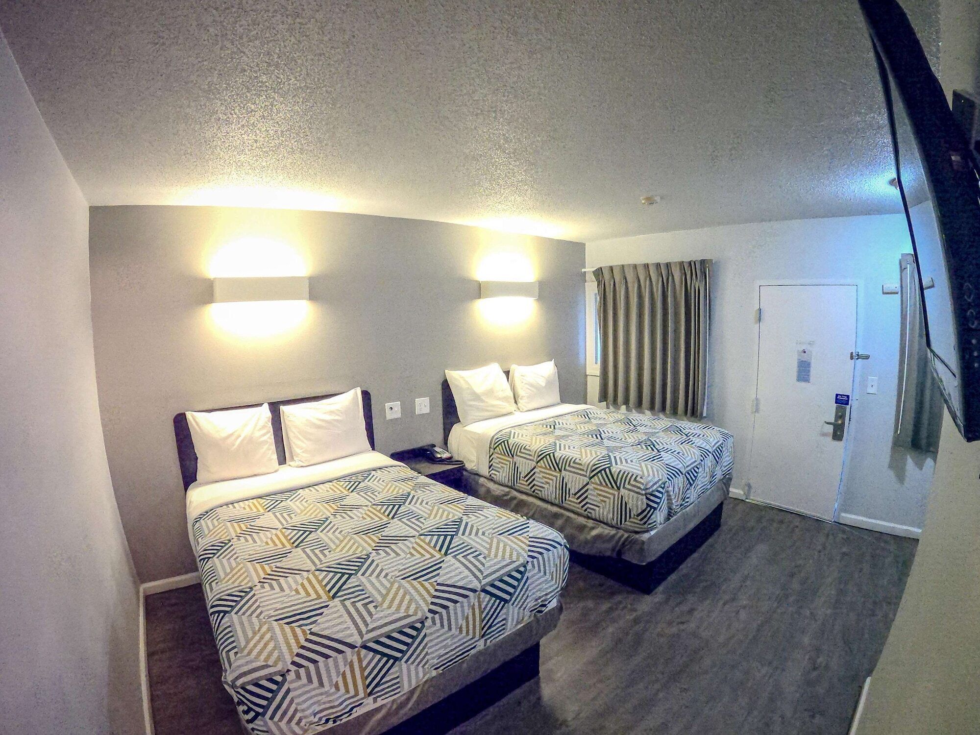 A hotel room with two beds, each with patterned bedding, a light-colored carpet, a door with a handle, and a wall-mounted light fixture.