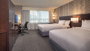 Habitación de hotel con dos camas, un escritorio, una silla y vistas a la ciudad a través de amplios ventanales.