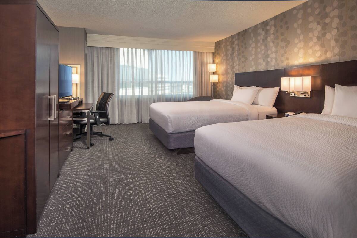 Quarto de hotel com duas camas, uma mesa, uma cadeira e vista para a cidade através de grandes janelas.