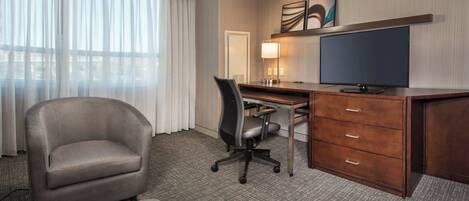 Une chambre d’hôtel avec un fauteuil gris, un bureau avec un ordinateur et un téléviseur à écran plat, ainsi qu’une grande fenêtre ornée de rideaux légers.