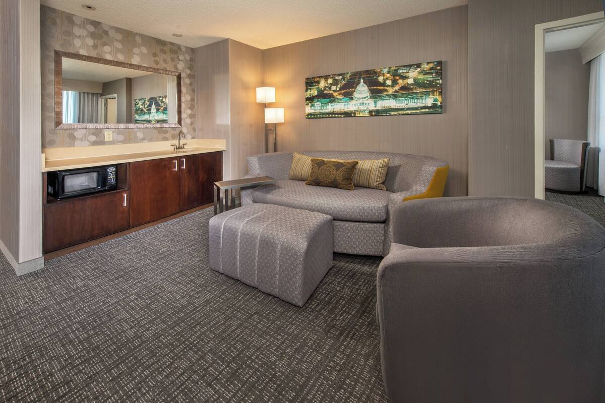 Quarto de hotel com cozinha compacta, sofá, cadeira, espelho grande e pintura pendurada na parede.