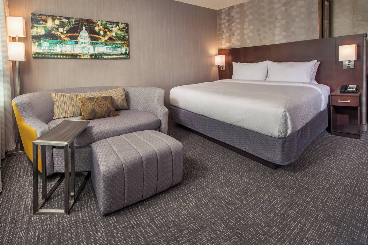 Quarto de hotel com uma cama grande, um sofá, uma mesa pequena e uma televisão na parede exibindo uma vista urbana.