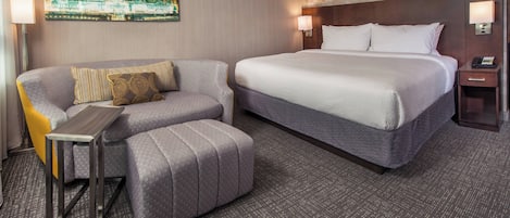 A hotel room with a large bed, a sofa, a small table, and a wall-mounted TV displaying a cityscape.