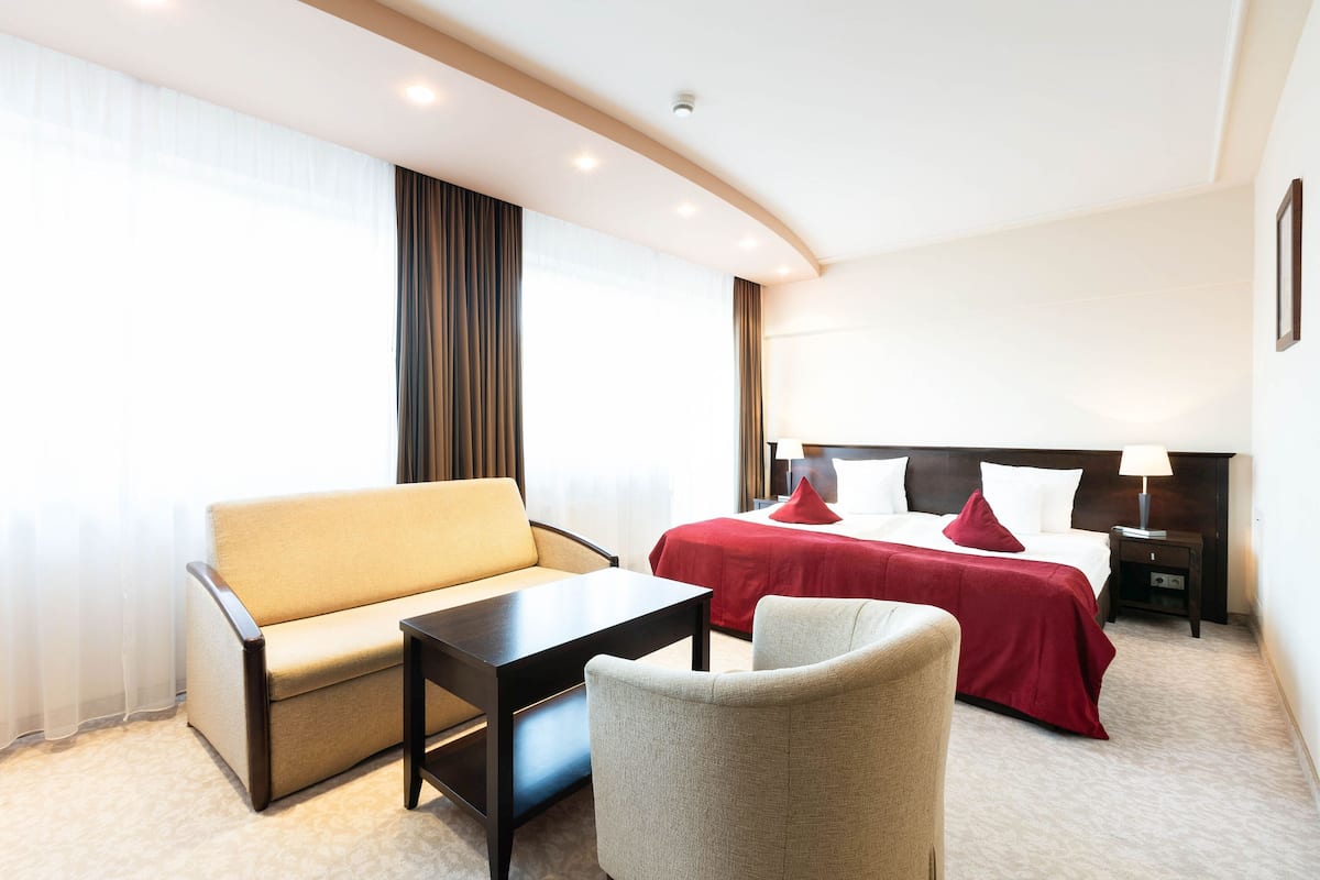 A hotel room with a bed, a sofa, a chair, a small table, and a nightstand with a lamp.