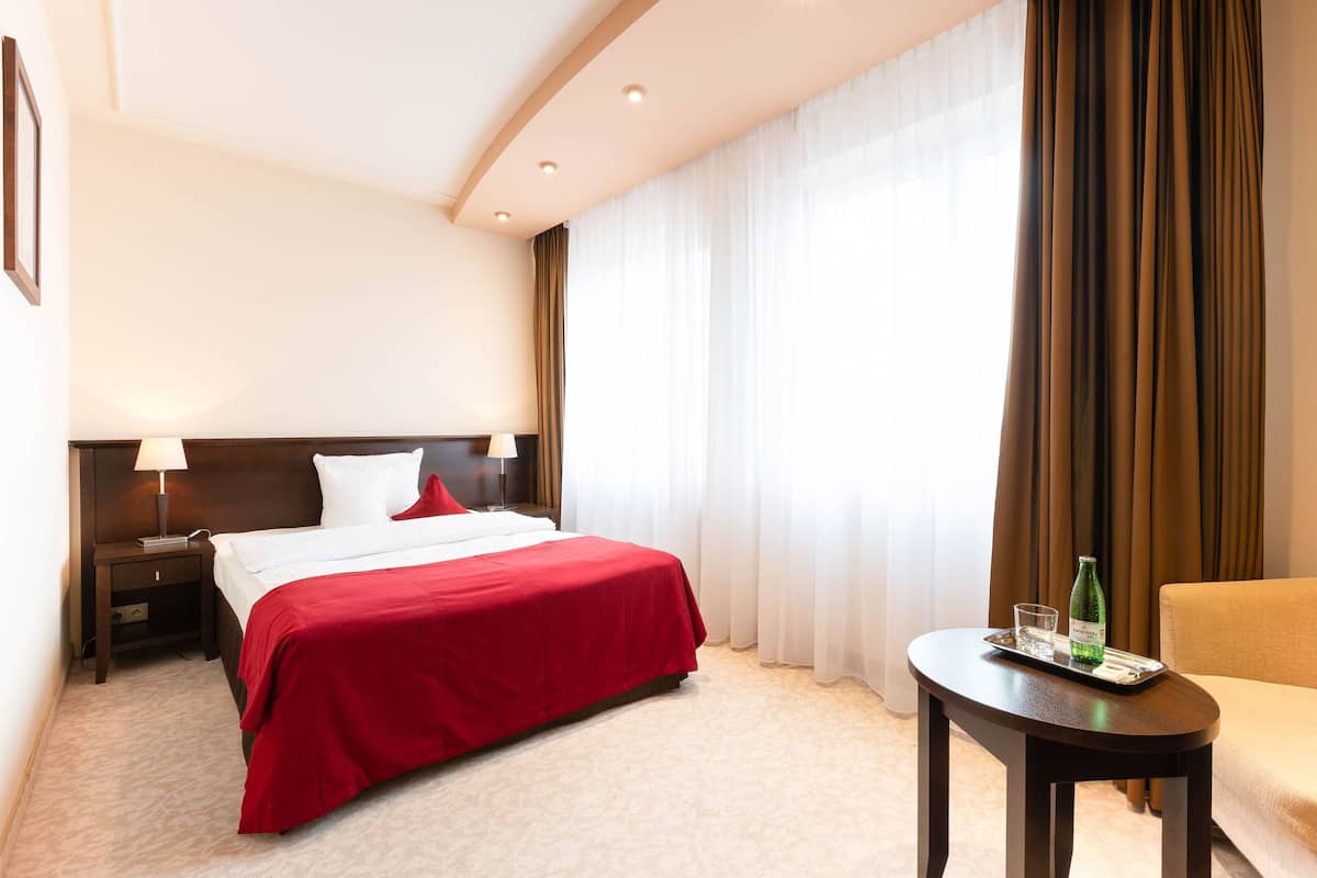 A hotel room with a bed, a nightstand, a lamp, a chair, a table with a bottle and glasses, and a window with curtains.