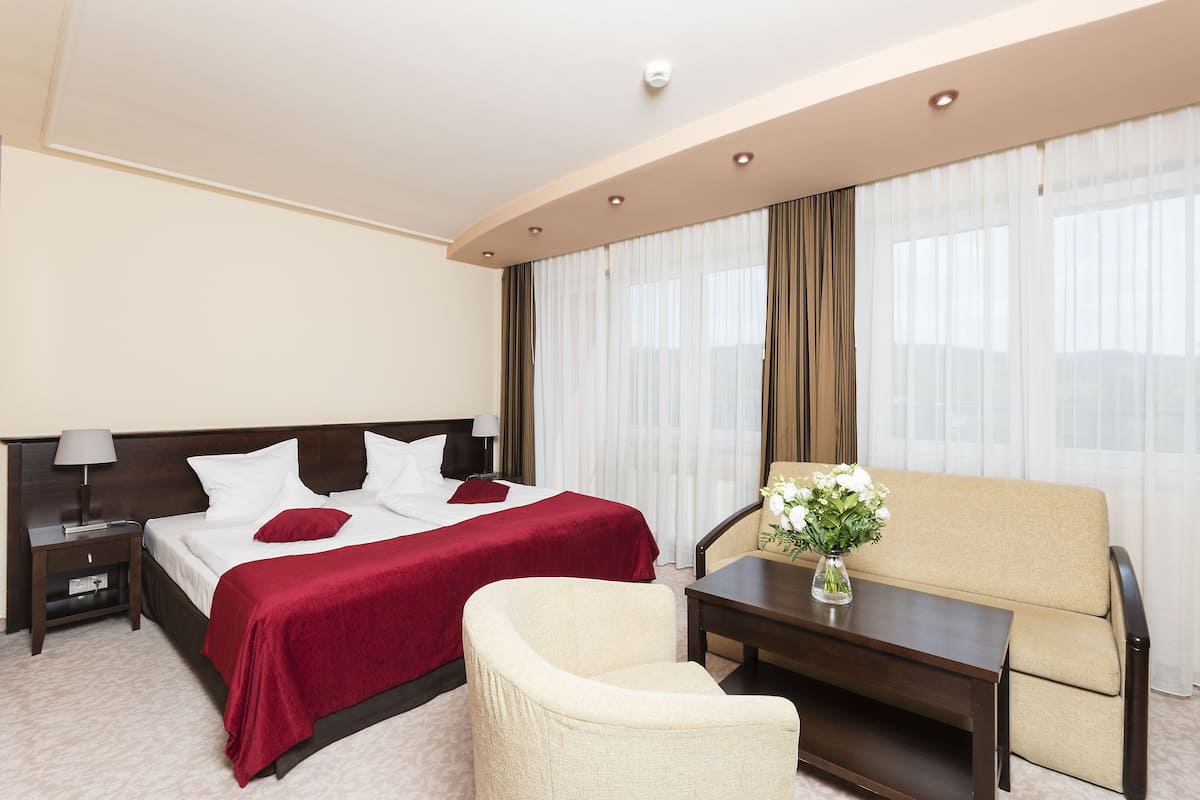 A hotel room with a large bed, a sofa, a chair, a nightstand, and a table with a vase of flowers.