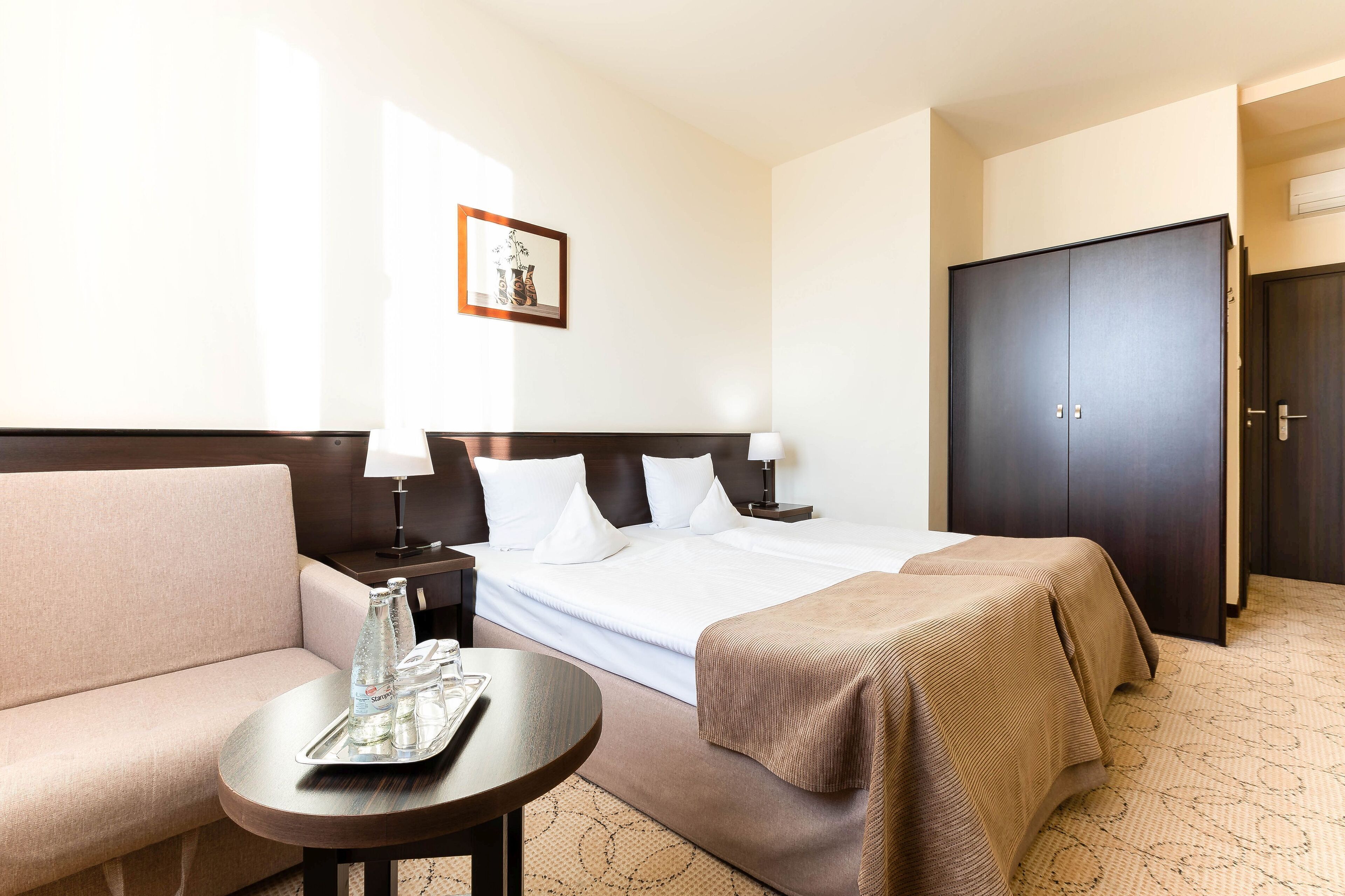 Ein Hotelzimmer mit einem Bett, einem Sofa, einem kleinen Tisch mit einem Tablett Wasserflaschen und einem dunklen Wandschrank.