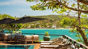 A poolside area with lounge chairs and umbrellas, overlooking a clear blue sea and a distant town.