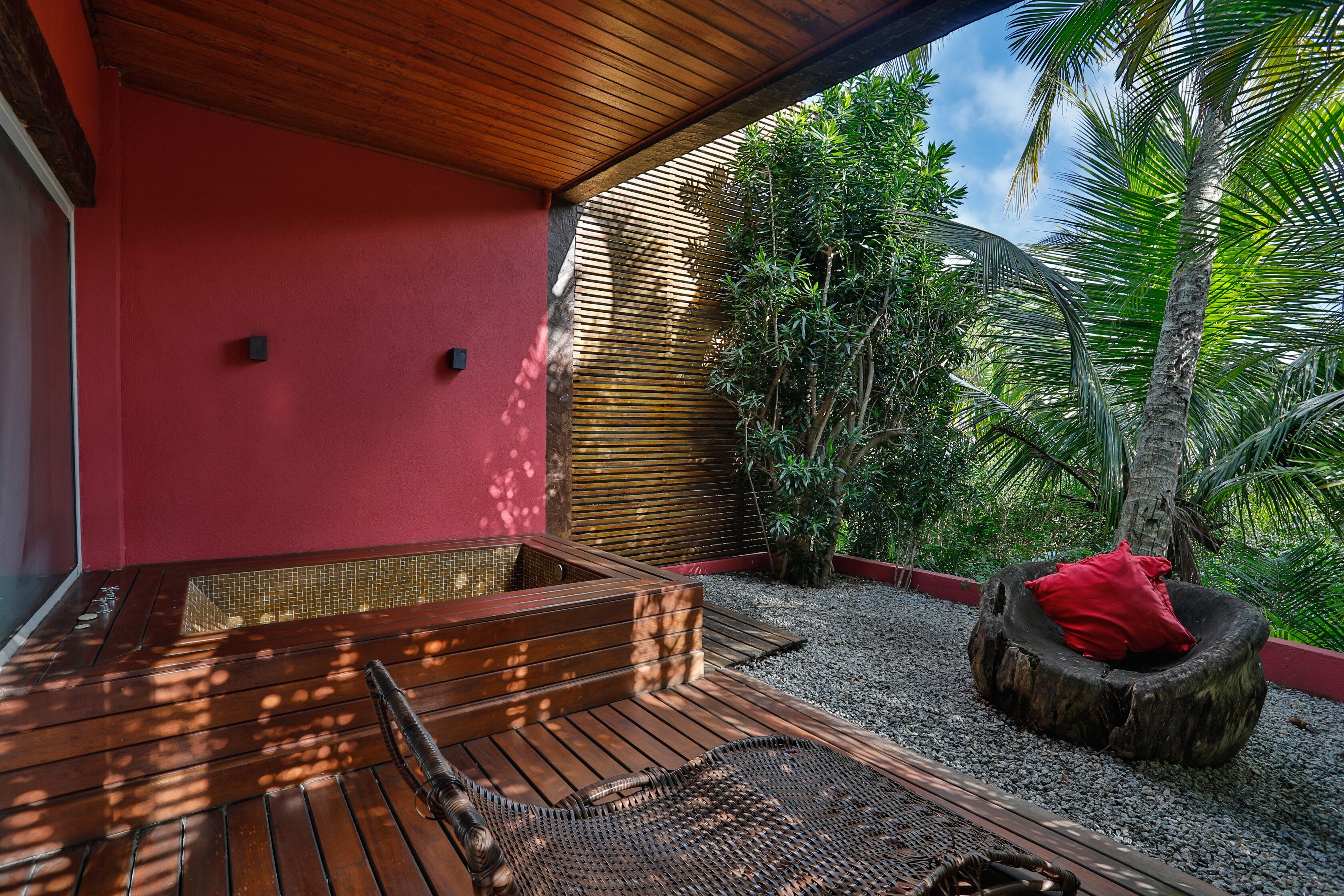 Una terraza de madera con un banco incorporado y un tapete tejido, rodeada por un muro rojo y plantas tropicales.