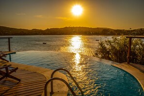 Une piscine dotée d’une terrasse en bois et de rampes en métal, surplombant une étendue d’eau où se trouvent des bateaux et une côte lointaine, le tout baigné par les couleurs du coucher de soleil.