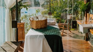 A room with a bed, a wooden table, a wicker basket, and a view of a body of water and greenery.