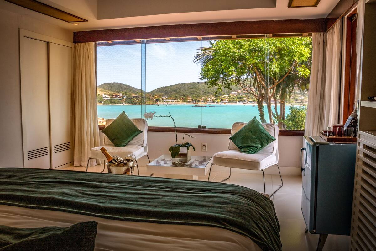 Ein Schlafzimmer mit einem großen Bett, zwei weißen Stühlen, einem kleinen Tisch und Blick auf Strand und Hügel.