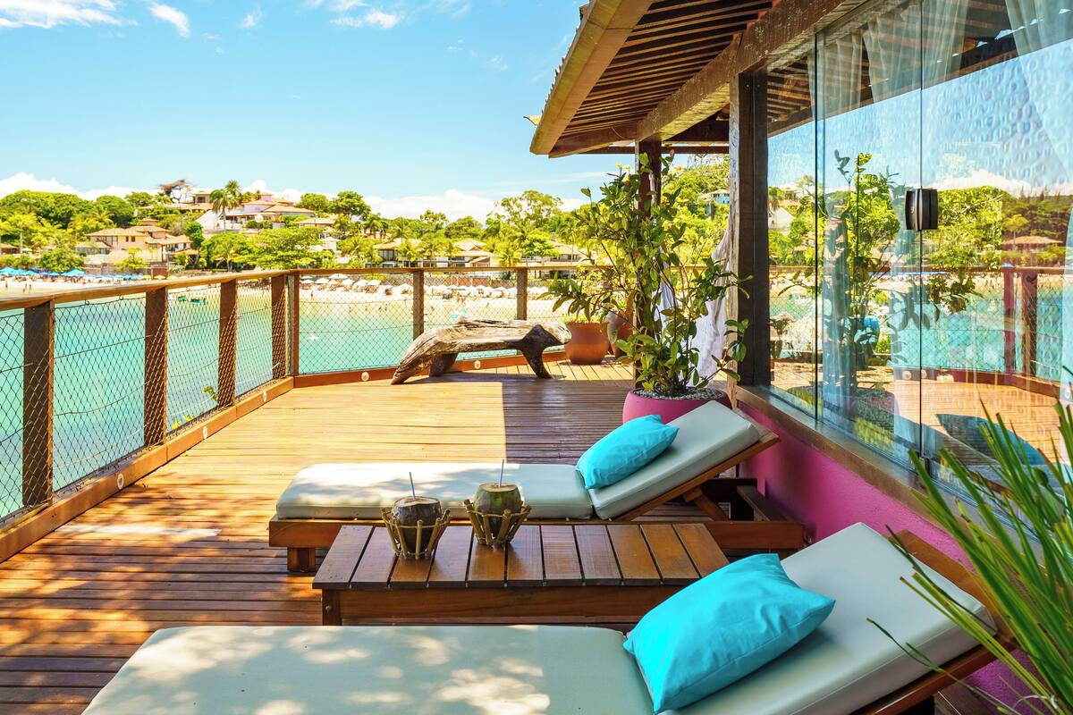 Eine Holzterrasse mit Liegestühlen und einer Glasreling mit Blick auf einen Strand.