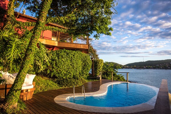 Une piscine dotée d’une terrasse en bois, entourée d’une végétation luxuriante et offrant une vue sur une étendue d’eau.