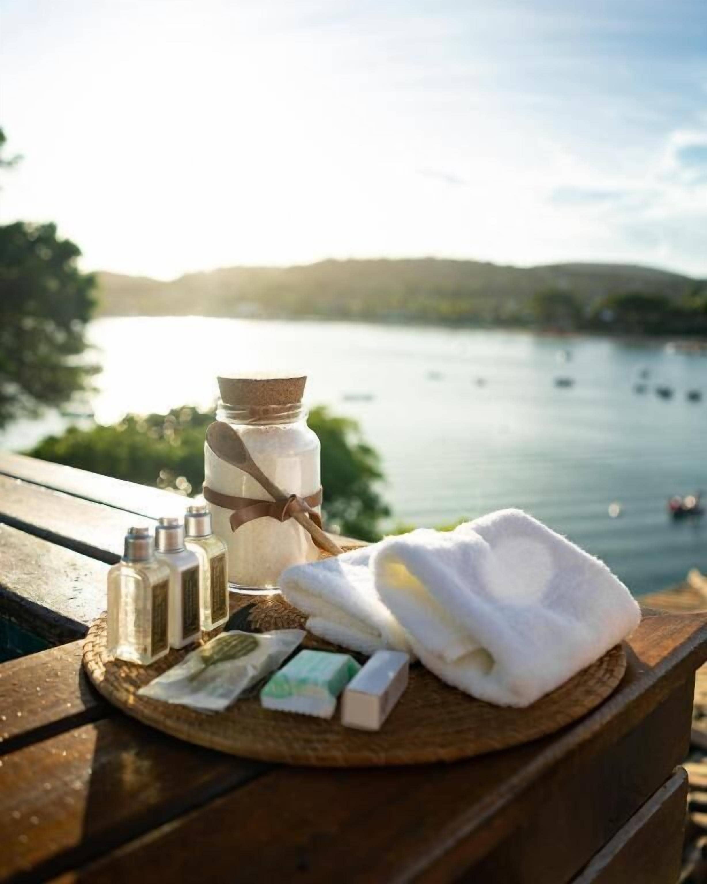 A wooden table with a bottle of lotion, a white towel, and some soap bars by a lakeside view.