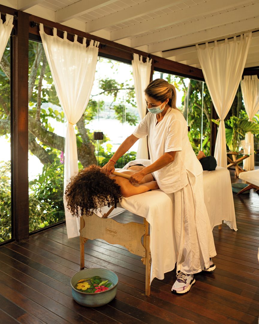 A massage therapy session in a room with wooden flooring and large windows, allowing natural light and views of greenery.