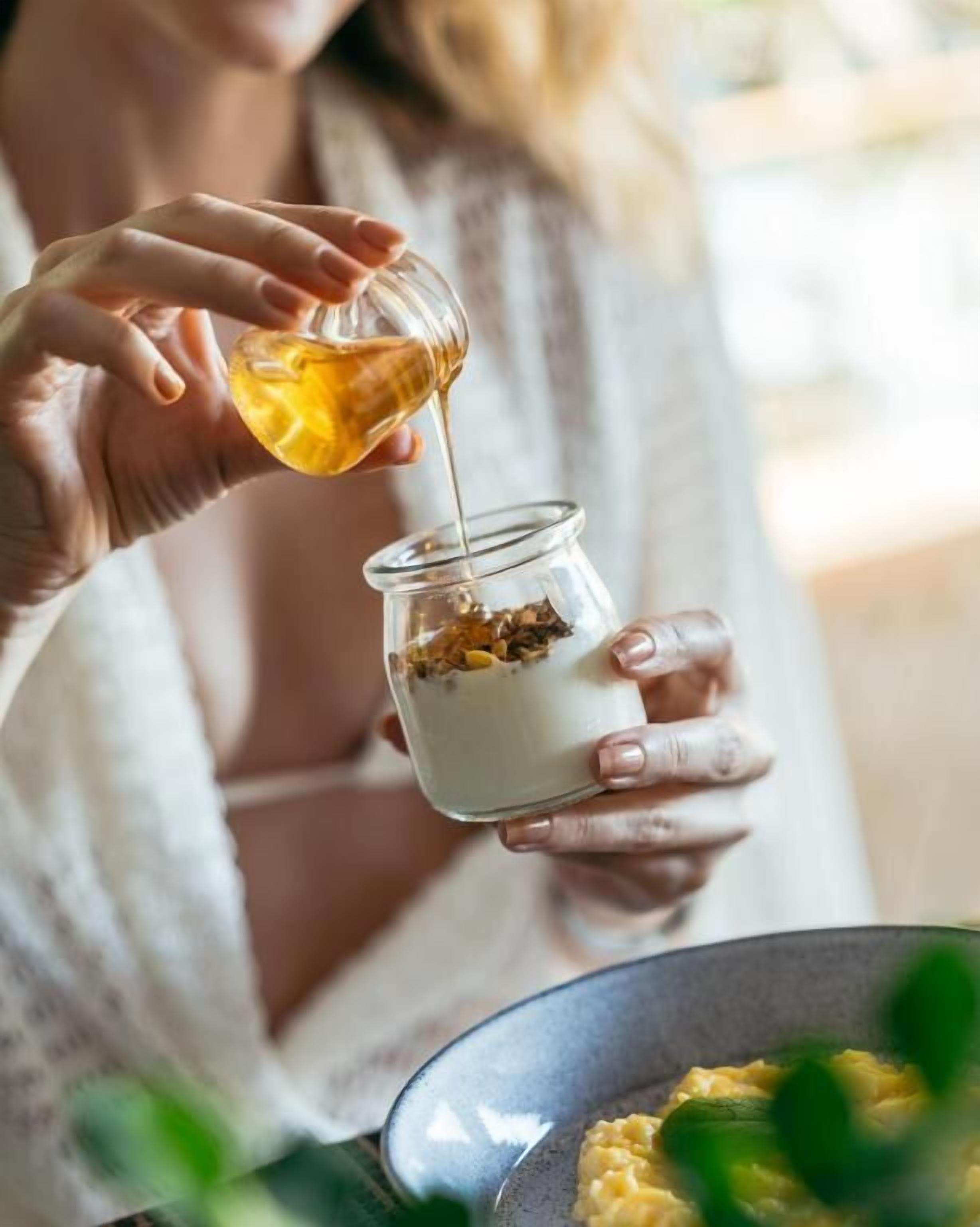 A person pouring honey into a jar containing a white substance, possibly yogurt or cream.