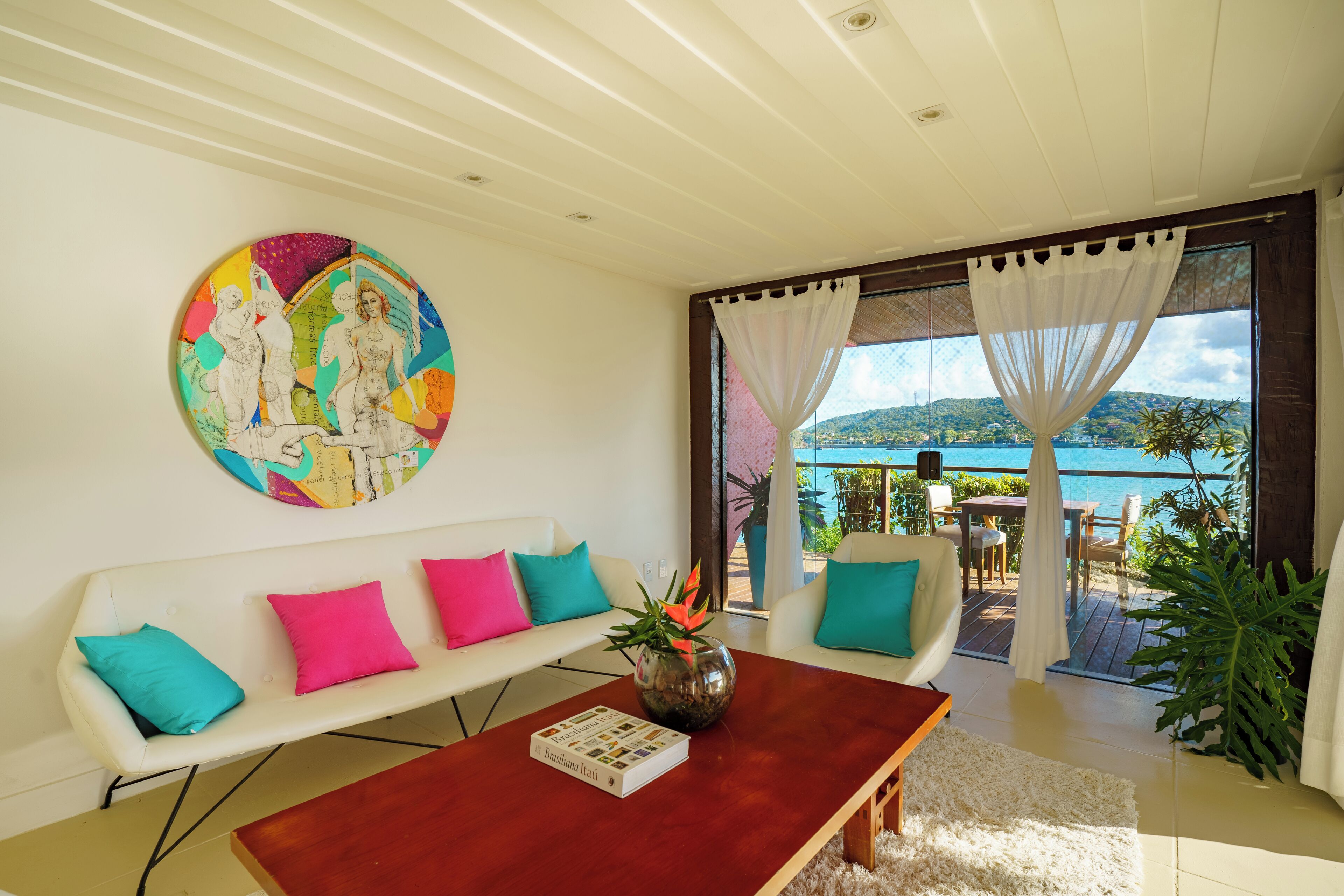 A modern living room with a white sofa, colorful cushions, a wooden coffee table, and a large window with a view of the sea.
