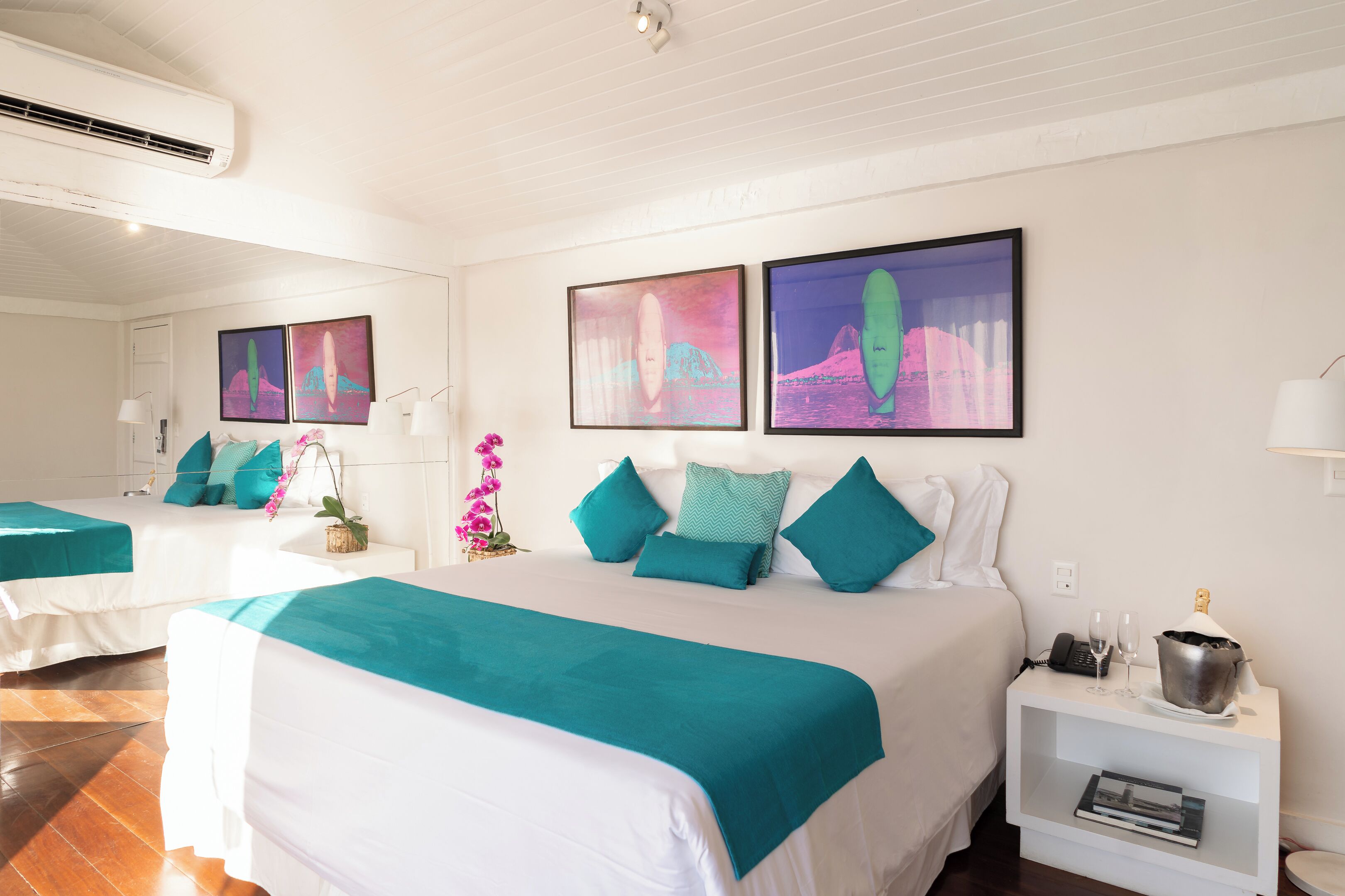 Habitación de hotel con dos camas, ropa de cama color aguamarina, una mesita de noche con teléfono y gafas, y dos obras de arte enmarcadas en la pared.