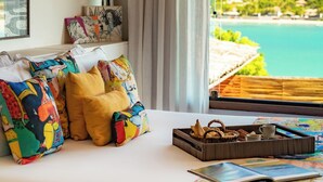 A bedroom with a bed, colorful pillows, a tray with snacks, and a view of a coastal landscape.