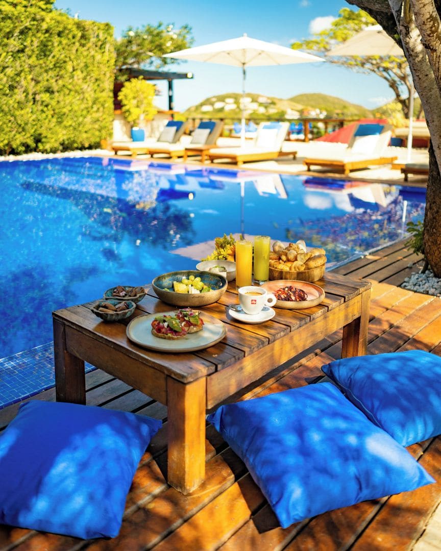 Une table en bois, dressée avec des aliments pour le petit-déjeuner, dans un cadre agrémenté d’une piscine.