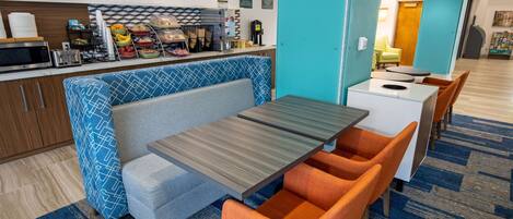 Un hall d’hôtel moderne doté d’un espace détente comprenant un banc aux motifs bleus et blancs, une table en bois avec des chaises orange, et un coin cuisine équipé de divers produits alimentaires et d’une machine à café.