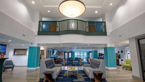 Un hall d’entrée spacieux avec un plafond haut, un espace de détente central avec des canapés, une table basse et un escalier menant à un niveau supérieur.