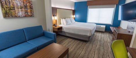 A hotel room with a bed, a blue sofa, a wooden coffee table, a desk with a chair, a flat-screen TV, and a painting on the wall.