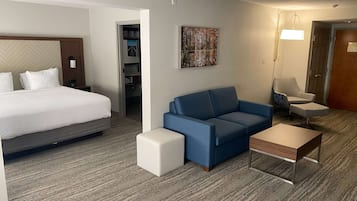 A hotel room with a bed, a blue sofa, a small white ottoman, a wooden side table, and a painting on the wall.