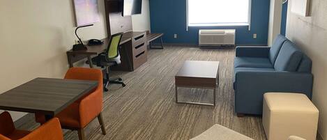 A hotel room with a desk, chair, sofa, ottoman, and a TV.