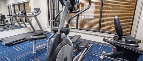 A hotel fitness center with cardio machines, a mirrored wall, and large windows with blinds.