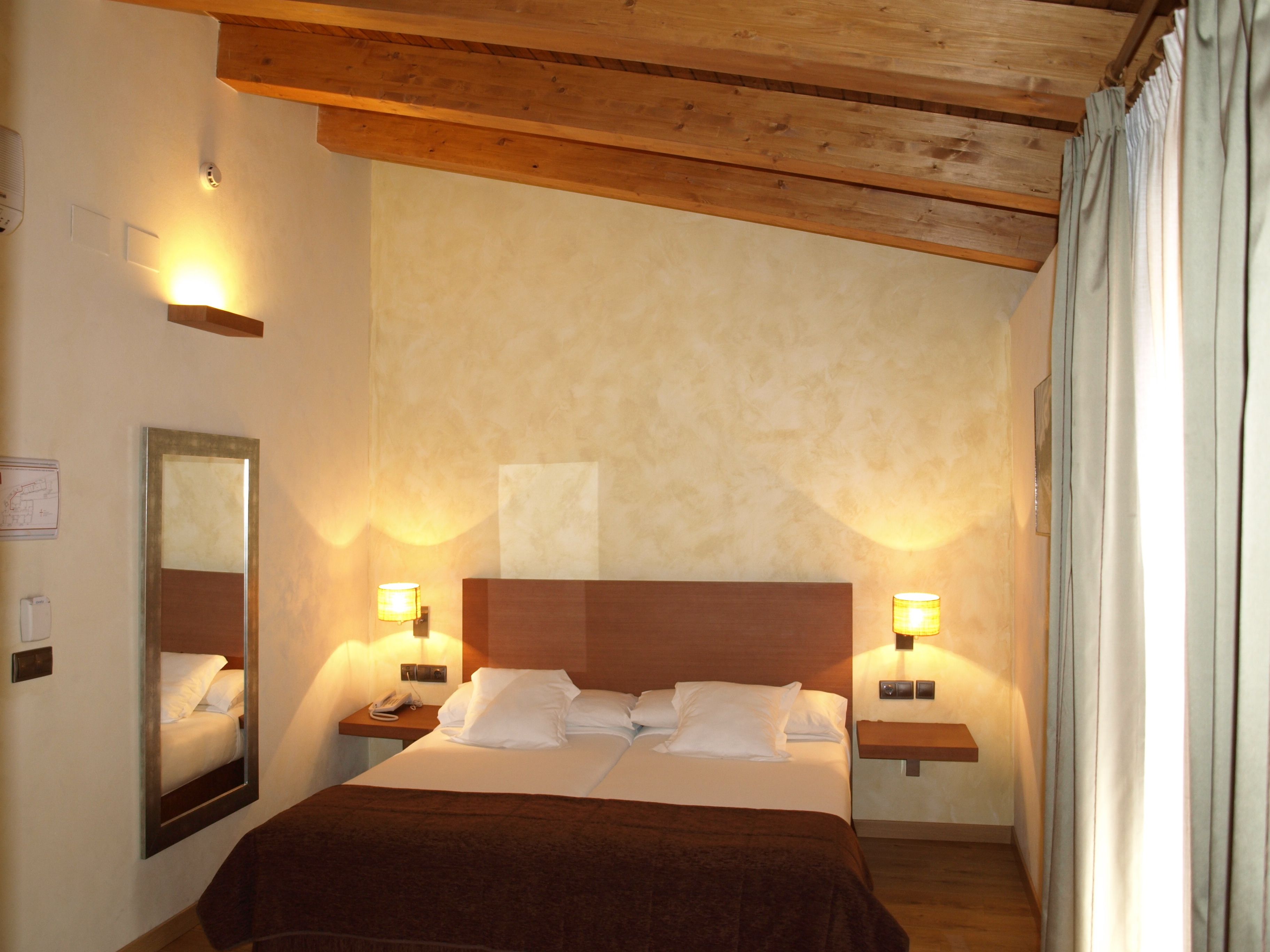 A hotel room with a wooden ceiling, a large bed with white linens, two bedside tables with lamps, a mirror on the wall, and a map on the left side.