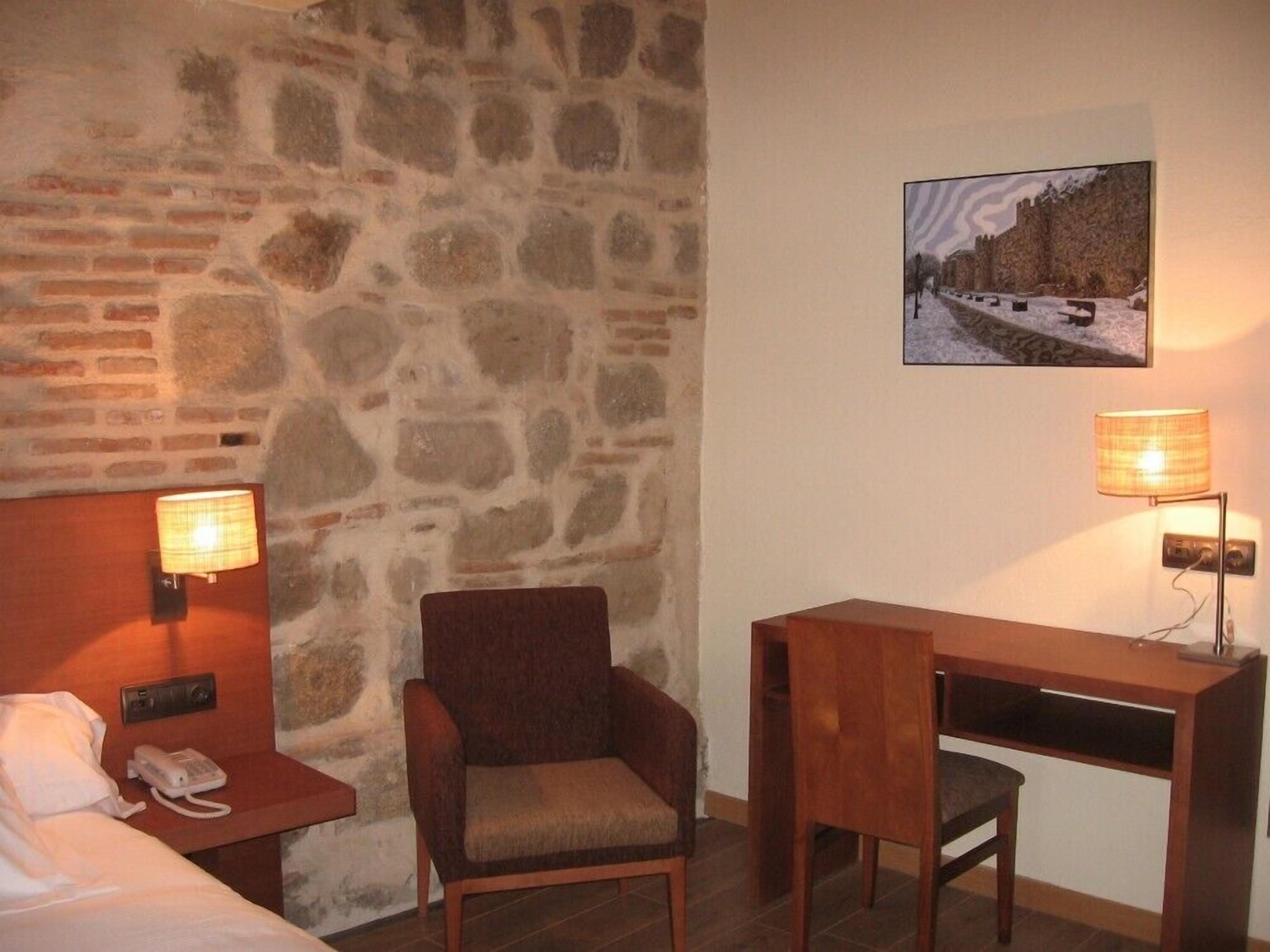 Habitación de hotel con pared de piedra, un escritorio de madera, una silla acolchada, una mesita de noche con lámpara y un teléfono.