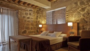 Una habitación con pared de piedra, una cama con ropa de cama blanca, un banco de madera, una silla y dos lámparas de noche.