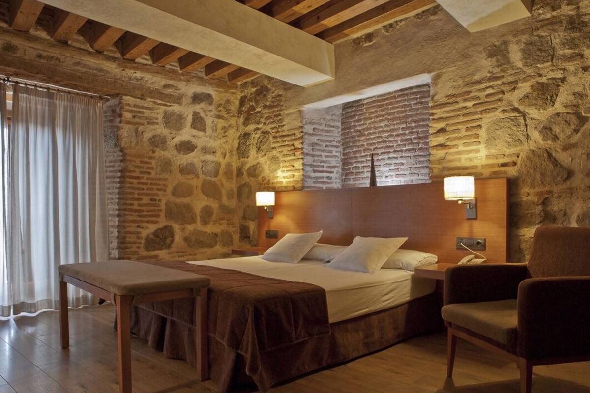 Une pièce avec un mur en pierre, un lit avec une literie blanche, un banc en bois, une chaise et deux lampes de chevet.