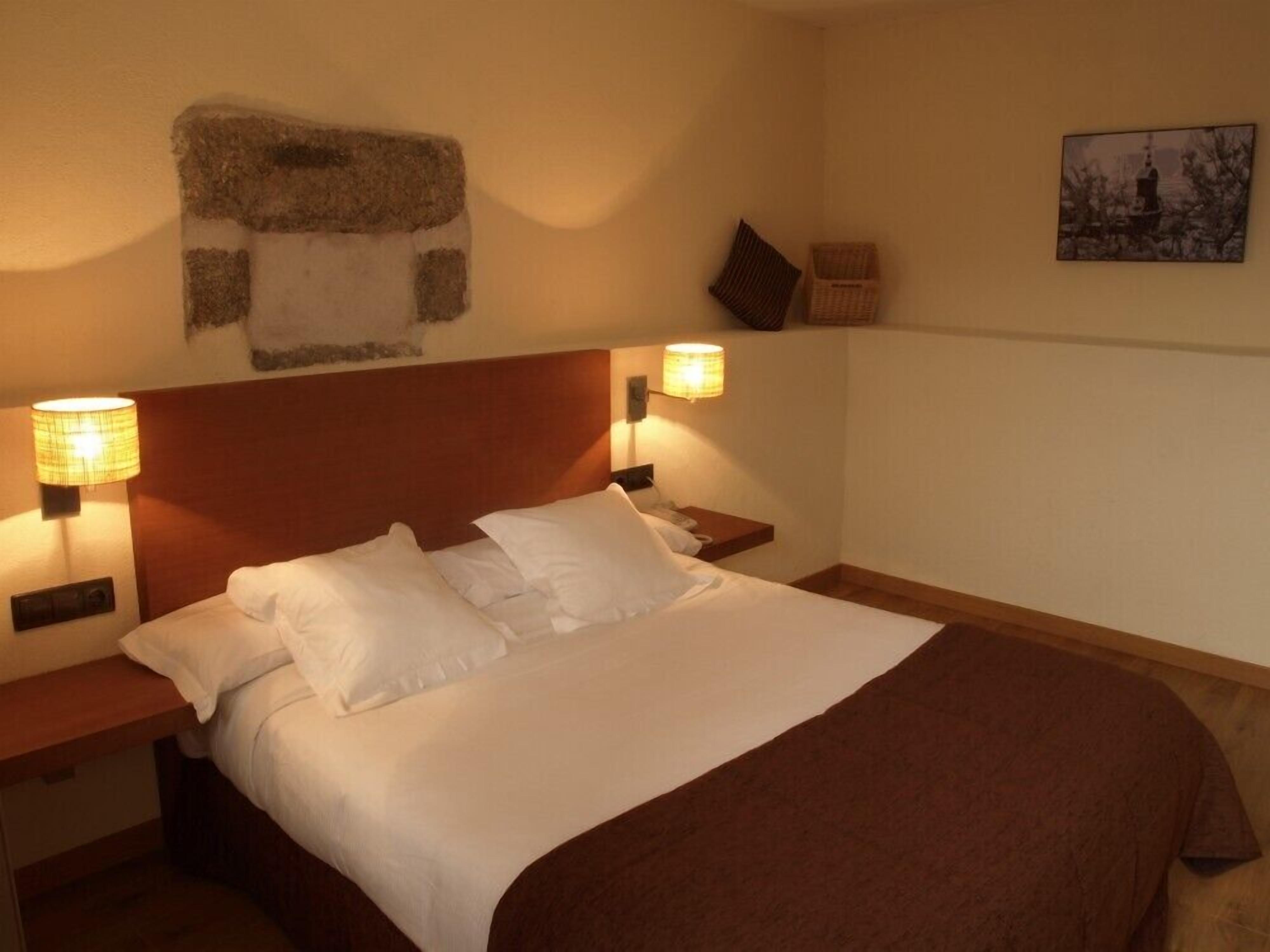 Habitación de hotel con una cama grande, dos mesitas de noche, un cabecero y dos lámparas fijadas a la pared.