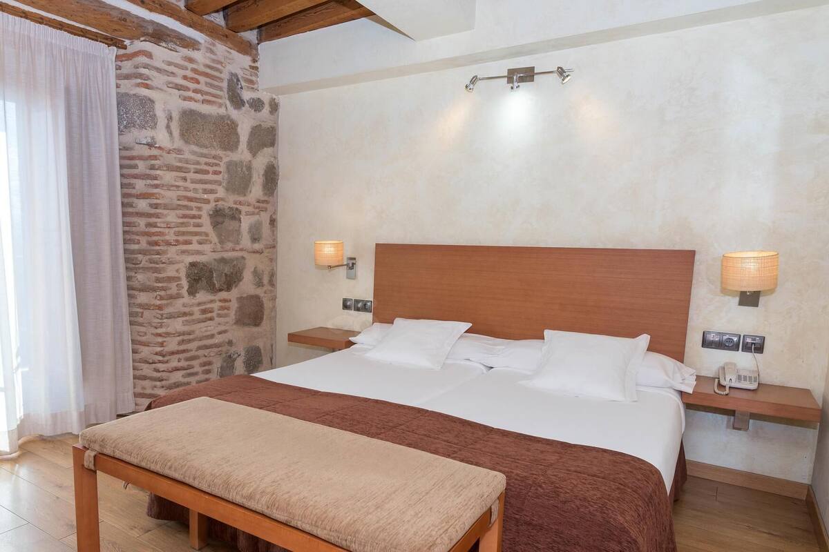 Une chambre à coucher avec un lit en bois, du linge de lit blanc, un banc, un mur en pierre et deux lampes de chevet.