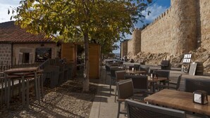 Un café al aire libre con paredes de piedra y mesas, rodeado de árboles y vistas a un edificio histórico de piedra.