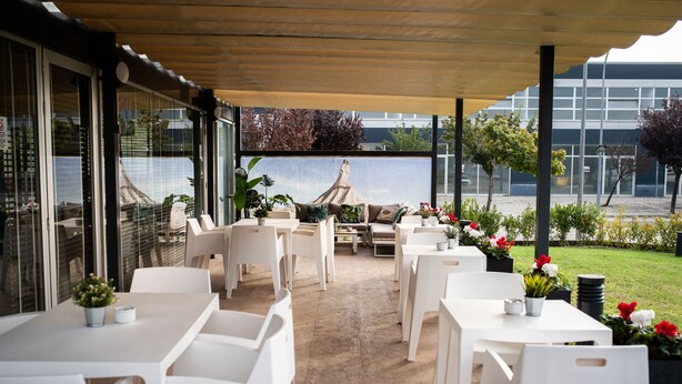 Un espace extérieur aménagé en terrasse, avec des tables et des chaises blanches, des plantes en pot et une vue sur un bâtiment et des arbres.