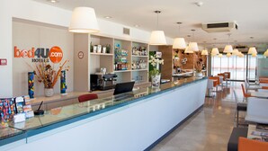 Un espace d’accueil d’hôtel avec un comptoir en verre, une machine à café et divers équipements, notamment une plante, un vase et un présentoir de cartes postales.