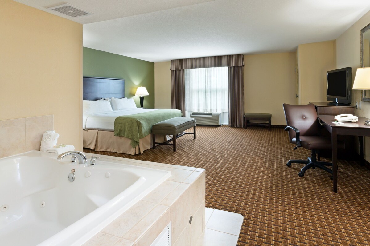 A hotel room with a large bathtub, a bed with a green headboard, a desk with a computer, and a television.