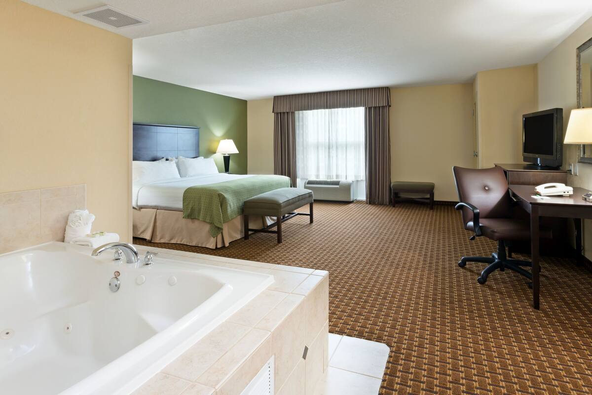 Ein Hotelzimmer mit einer großen Badewanne, einem Bett mit grüner Bettdecke, einem Schreibtisch mit Computer und einem Fernseher.