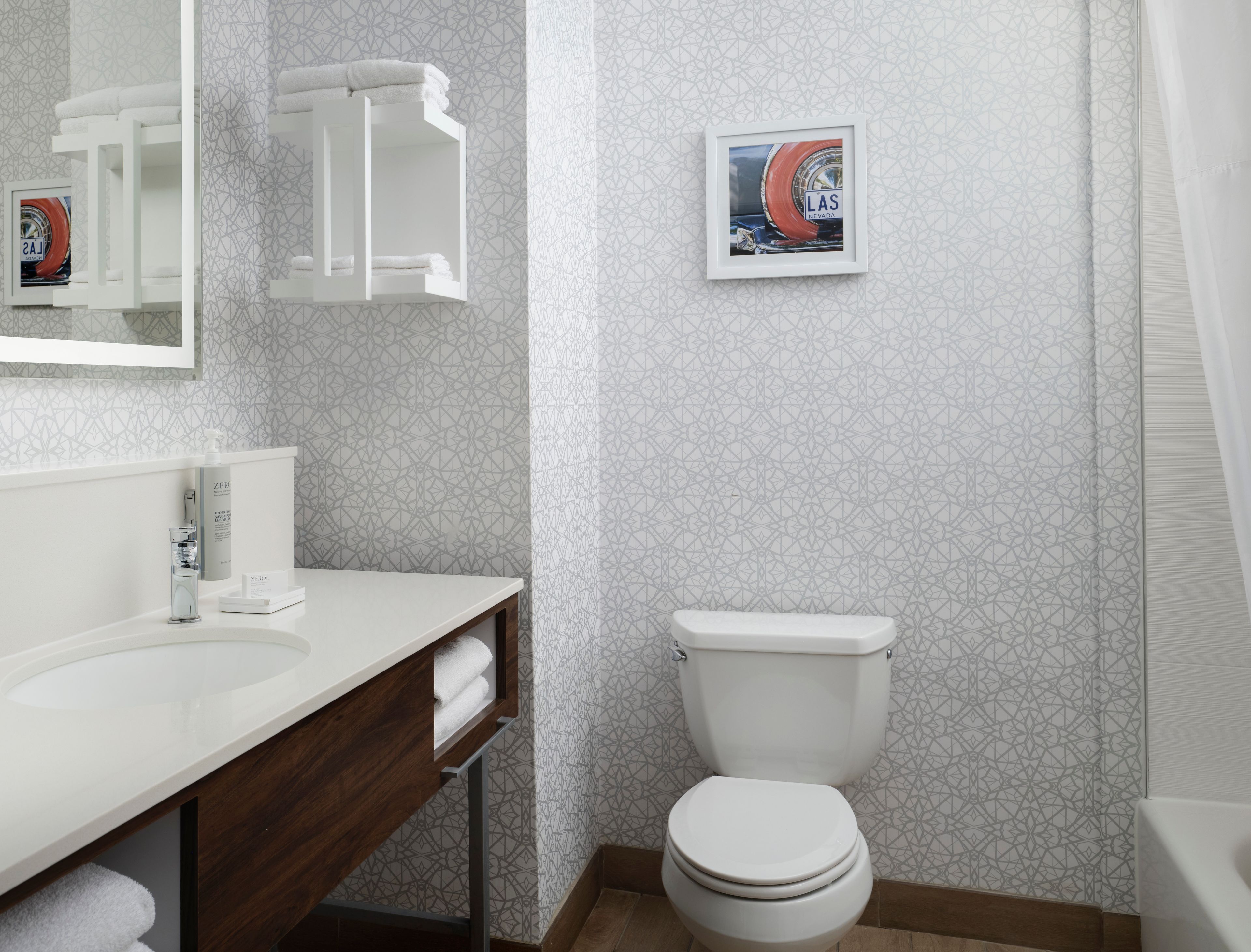 Une salle de bain moderne avec un lavabo blanc, un miroir et des toilettes. Un tableau encadré est accroché au mur, et il y a un porte-serviettes.