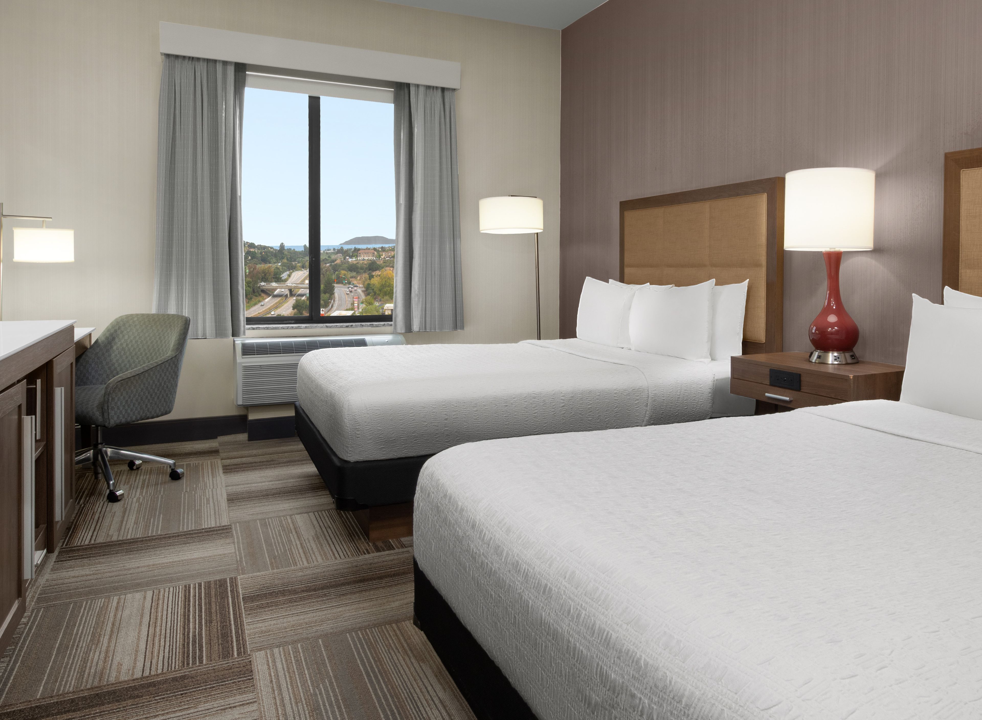 Habitación de hotel con dos camas, un escritorio, una silla, un ventanal con vistas y dos lámparas.