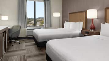 Habitación de hotel con dos camas, un escritorio, una silla, un ventanal con vistas y dos lámparas.