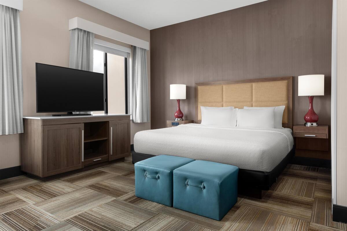 A hotel room with a large bed, a television, two bedside lamps, and a small blue ottoman.