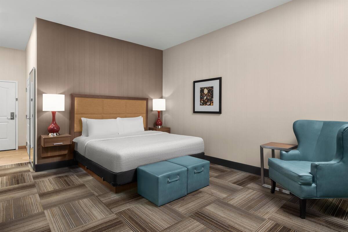 A hotel room with a large bed, two bedside tables with lamps, a blue armchair, and a small table.