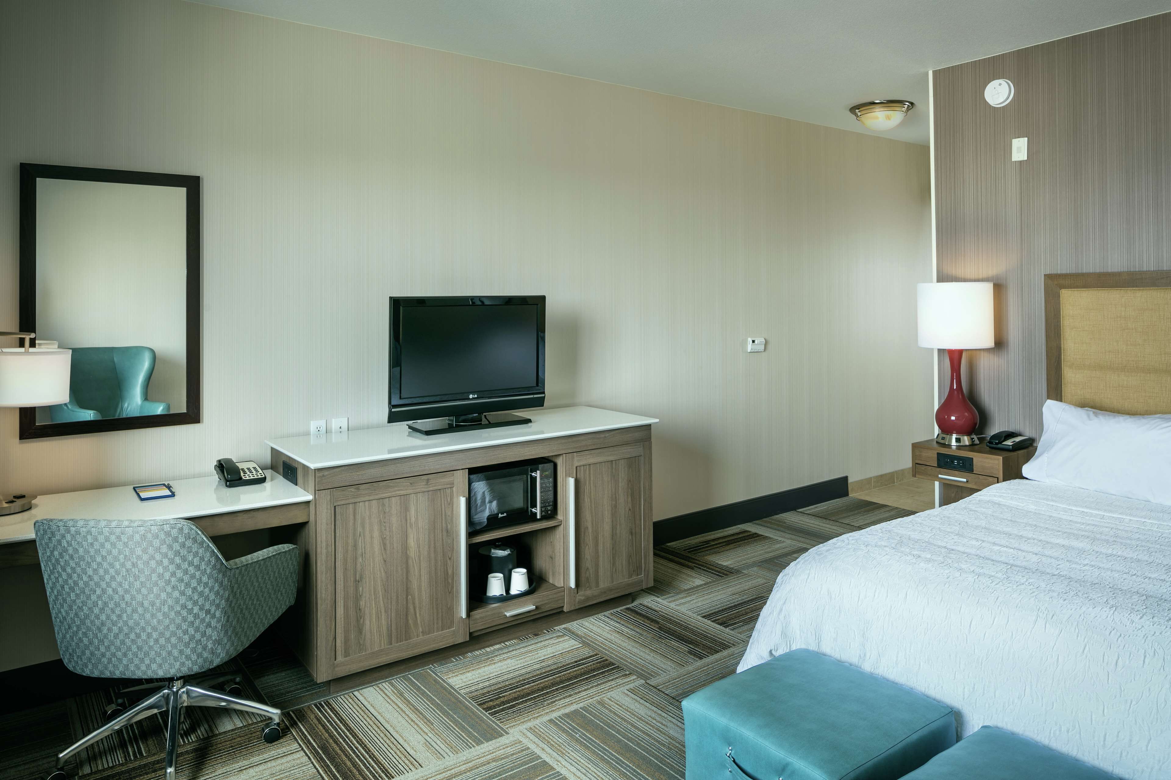 A hotel room with a bed, a desk with a television, a chair, a lamp, and a microwave.