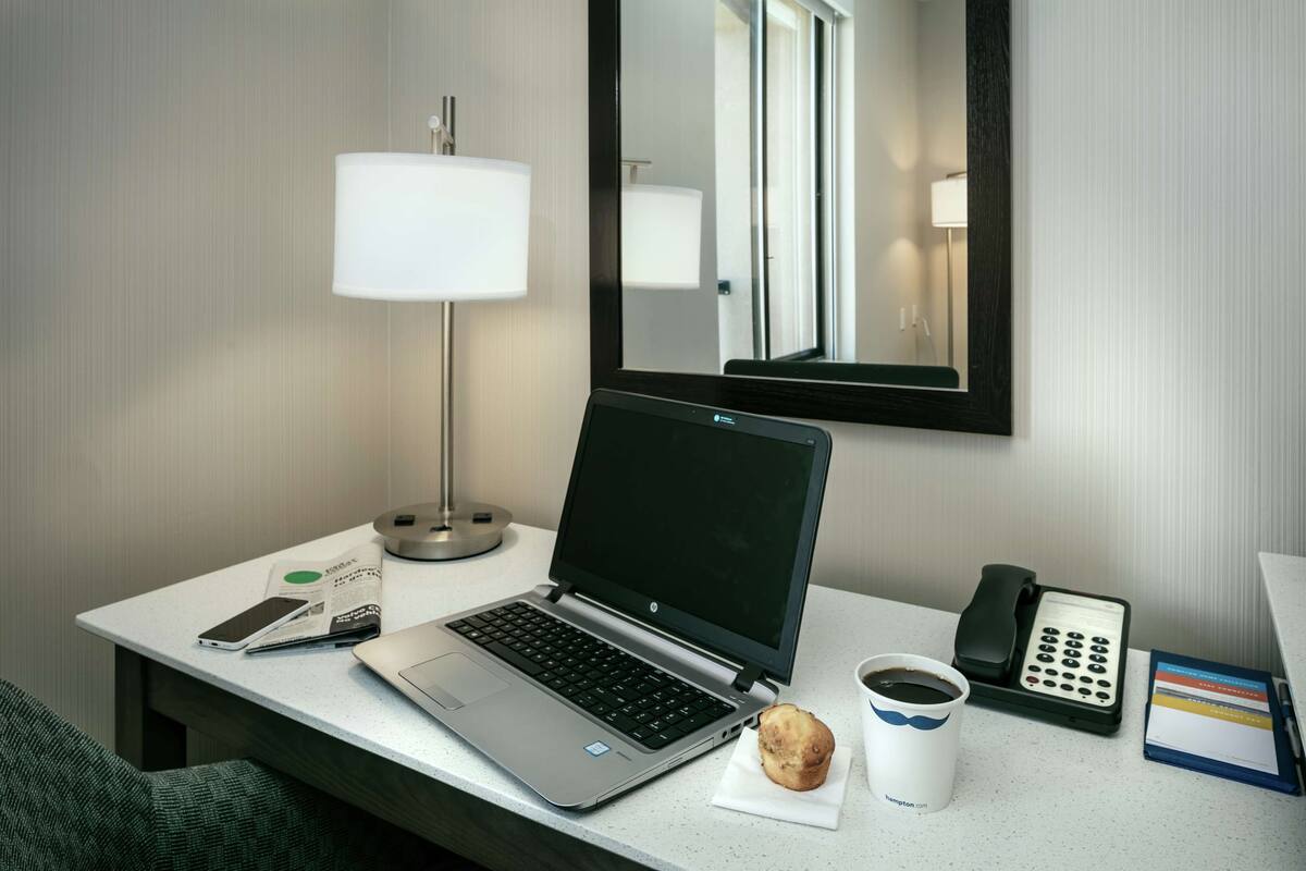 A hotel room desk with a laptop, a mobile phone, a coffee cup, a muffin, a telephone, and a notepad.