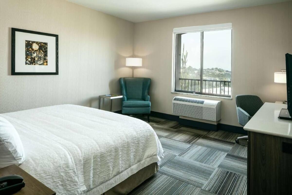A hotel room with a bed, a desk, a chair, a window with a view, and an air conditioning unit.